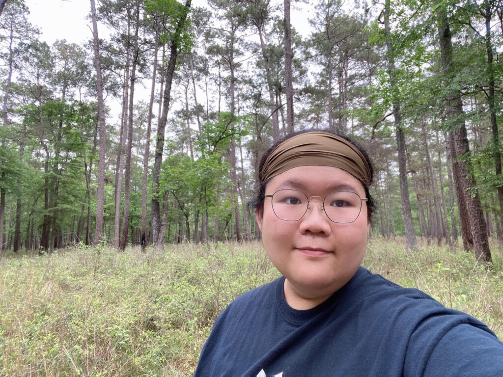 Selfie of person with hair pulled back, wearing a brown headband. Person is stanidng outside in front of an outcropping of tall trees