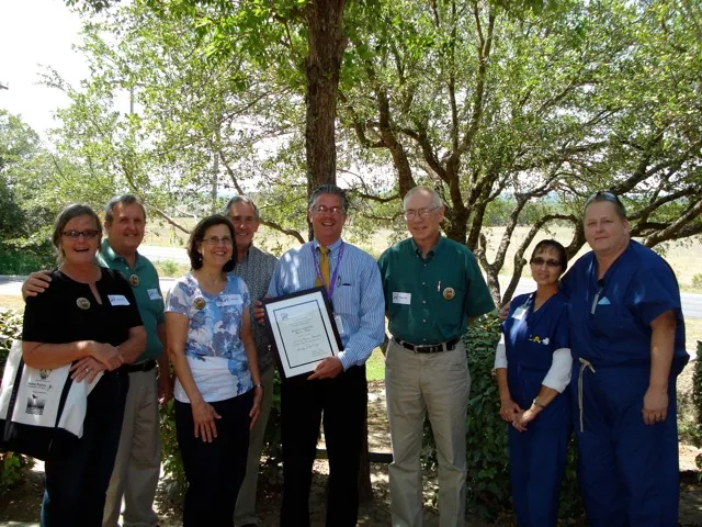 Group of people standing in front of a tree. The person in the middle is holding a framed certificate.